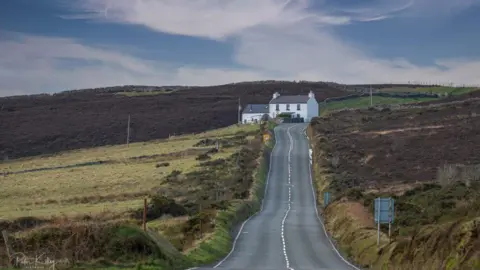 MANX SCENES A long winding road with an embankment on the right, and green hills on the left, a white cottage is on the right of the road at the top. 