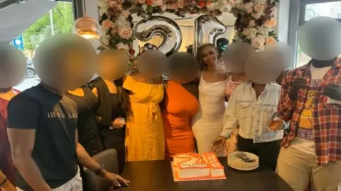 A group of 11 young people stand close together next to a cake in the shape of the number 21. All their faces are blurred apart from that of a young woman smiling in the middle.
