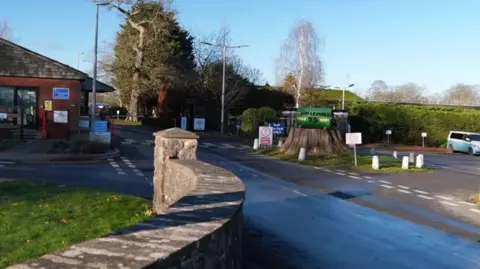 The road leading towards the entrance to HMP Leyhill on a sunny day. At the front of the image is a low stone wall. There is a large tree stump just right of the centre which has a green sign on it saying 'HMP Leyhill 75 1946 2021'. There are trees in the background, a small redbrick building to the left of the image and a blue and a white camper van parked to the right. 