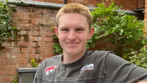A photo of a man sitting on a bench smiling into the camera. He has got short orange bleached hair and it is very short. You can only see his head and shoulders. He has got a gray top on which has stitching on it with BRONCO written. 