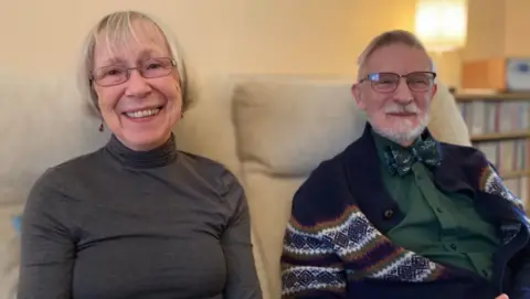 Eve and Peter Beyfus smiling while sitting on couch in home. They are both wearing glasses, Eva is wearing a grey top and Peter is wearing a bowtie with a green shirt. 