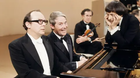 Eric Morecambe, Ernie Wise and Andre Previn on 'The Morecambe and Wise Christmas Show' in 1971. Eric and Ernie sit at a piano in black tuxedos while Andre Previn leans on the piano to the right with his head in his hands at he stares at Eric