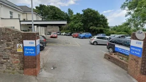 The Chadwell Centre in Paignton. The car park outside the white and cream-coloured building is filled with vehicles. A brick wall is on the perimeter of the car park. There are blue and white NHS signs saying the car park is private. Trees are dotted around the opposite side of the car park.