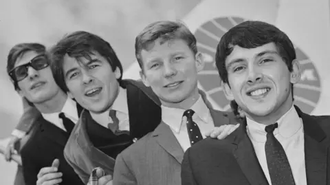 A black and white photo of The Searchers. From left to right, drummer Chris Curtis, guitarists Mike Pender and John McNally and bass player Frank Allen pose on the steps to a plane at London airport. They are all wearing suits, shirts and ties. 