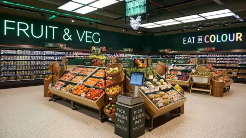 The fruit and vegetable section of a Marks and Spencer store. There are two islands with brightly coloured displays of fruit and vegetables, and a set of self-service weighing scales in the foreground.