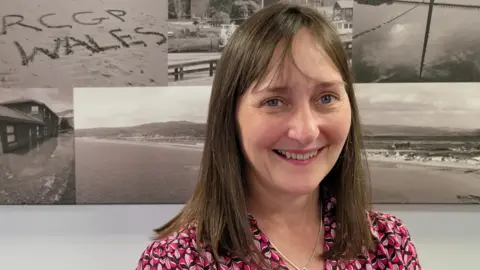 Dr Rowena Christmas has brown shoulder length hair and a partial fringe. She has blue eyes and is smiling at the camera. She is wearing a patterned shirt with different shades of pink and is stood in front of some black and white beach photos