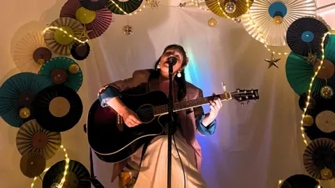 CHIp A woman sat a stool playing a guitar and singing. the stage is surrounded by decorations.