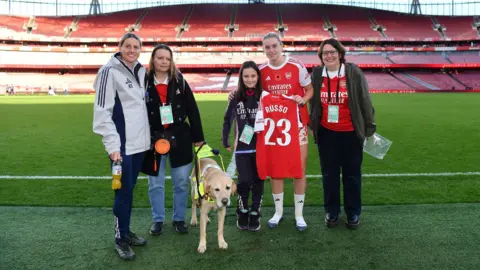 Blind Veterans UK A group of people stood on an artificial pitch with a guide dog between them. One is a footballer who holds a strip which says 'Russo 23'