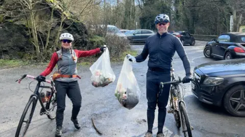 A man and a woman wearing cycling clothes are standing beside bicycles in a car park, each holding a large white bag containing litter