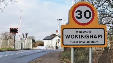 Road sign to the right of a road heading over a hill and level crossing with homes beyond.