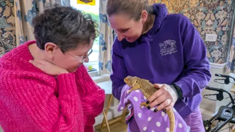 A woman in a purple jumper featuring a white logo holds an iguana in a spotted cloth. A woman wearing a pink jumper is standing next to her and is looking at the lizard.