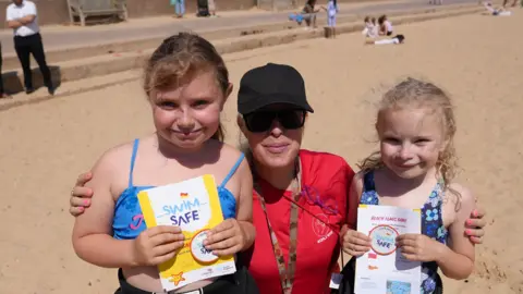 Qays Najm/BBC Rosa (left), mum Rosie and Flora (right) standing on the beach. The young girls are holding swim safe certificates. 