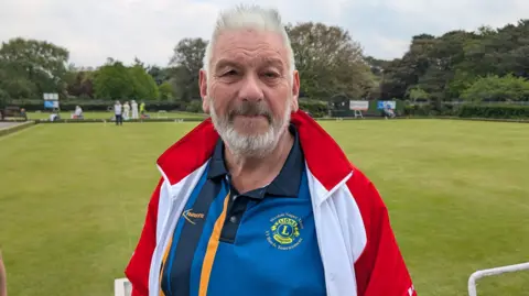 An older man stands in front of a bowling green. There are people playing bowls behind him, wearing white. He is wearing a blue bowls shirt with yellow and black stripes, and a red and white jacket. He has grey hair and facial hair.