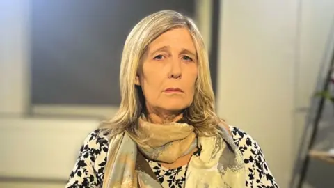 BBC Vanessa is looking at the camera. She is wearing a black and white patterned long sleeve top and a brown, blue and cream scarf. She has shoulder length blonde hair. Behind her, there are shelves with plants on them and a window with blinds shut.