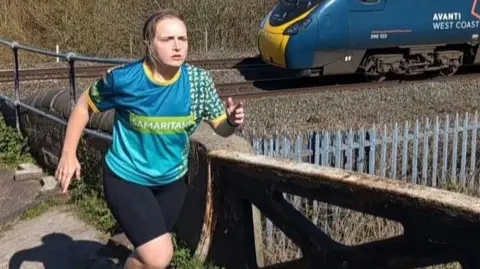 A woman wearing a blue running top which reads 'Samaritans' on it, black shorts and blue trainers, with her blonde hair tied back, runs across a bridge. A blue train is on a track underneath the bridge, which reads 'Avanti West Coast' on the side of it. It is a sunny day.