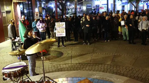 East Bay Times Via Getty Images Protesters have formed a semi-circle next to a woman who is speaking into a microphone. She is standing next to a drum kit.