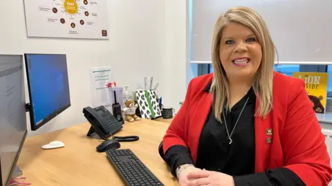 A woman with long blonde hair, red blazer, black top and wearing a silver necklace, smiles at the camera. She is sat at a light brown desk with a black monitor and a keyboard in front of her. There is also a telephone, walkie talkie, small teddy bear and some stationary on the desk. There is a window with a white blind half down behind her. 