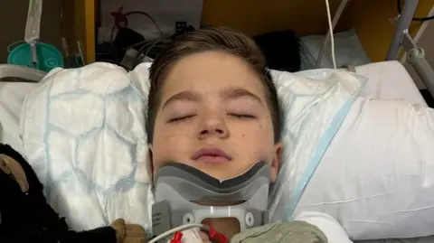 A young boy laying in a hospital bed with his eyes closed and his neck in a brace