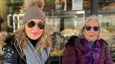 Joanne Baker and her mum sitting outside a bakery in Keyworth. Joanne, left, is a white woman with medium-length blonde hair wearing a beige woolly hat with a large bobble and a black padded coat and grey scarf. Joanne's mum is wearing a purple padded coat with a pink and purpled check scarf. 