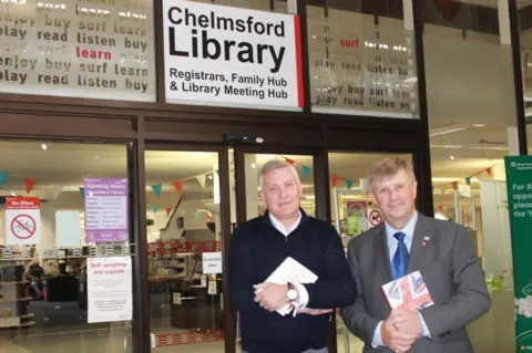 Essex County Council Russell Quirk, wearing a dark-coloured fleece, and Peter Harris in a grey suit and blue tie, smile directly at the camera. They are holding books and standing outside the glass sliding doors of Chelmsford Library.