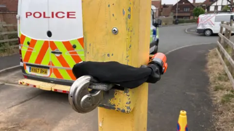 A black bicycle lock is wrapped around a yellow metal gate post. A police van is parked in front of the entrance. 