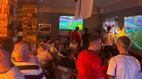 Jason Cleary People gathered inside a pub watching a football match on big TV screens on the wall. England flags are visible dangling from the ceiling.