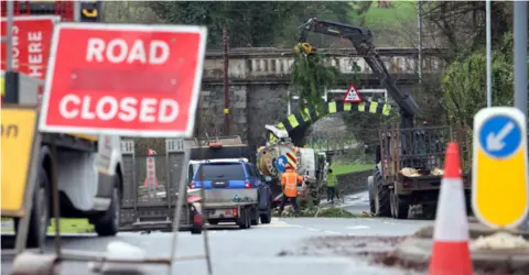 Pacemaker A shot of works being carried out on a road where branches of a tree have fell on the road. There is a red 'road closed' sign and workers with a blue pick-up truck, a tractor and trailer.