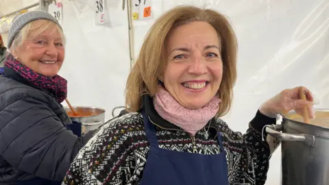 Elaine Ellis is to the left and is wearing a blue scarf with pink dots and a black coat. She has white hair and has a knitted hat on as well. Angela is closer to the camera and is wearing a Christmas jumper with a pink scarf. She has light brown hair which comes down to her shoulders. They are both stirring soup.