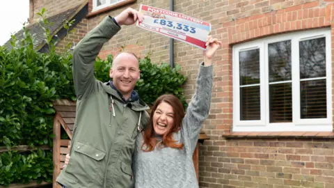A couple stand outside a house smiling while holding up their £83,333 winning lottery ticket 