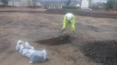 Leeds City Council An archaeologist wearing yellow hi-vis overalls and a white hard hat digs a hole in open ground, with white bags of rubble to the side.