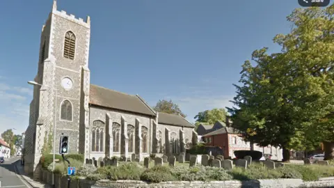 Google A medieval church in a town centre, with gravestones in the foreground, a tree to the right, and a set of traffic lights and a road to the left 