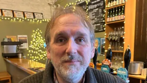 Head and shoulders image of Jamie Delap from the Society of Independent Brewers and Associates. He's standing in front of a bar with beer taps and a coffee machine the background. He has grey hair and a short grey beard and moustache and is wearing a brown, ribbed jumper.