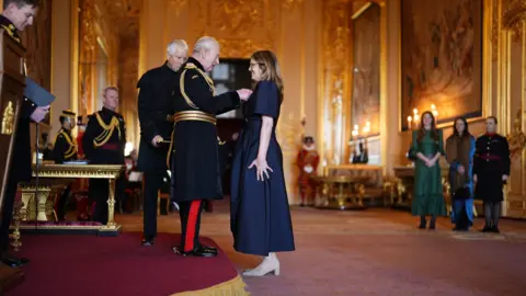 Jordan Pettitt/PA Wire A woman wearing a navy dress with balloon sleeves receives an award from the King in a large room at a castle. Other people in uniform can be seen in the background.