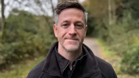 John Martin John Martin's hair is dark on top and grey at the sides. He is wearing a coat and fleece and there are trees in the background 