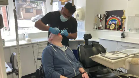 A man in a blue hoodie has his mouth open and is being examined by a dentist, wearing a mask and gloves and a black top. On the wall of the examination room is a sign with 'save the NHS' and a number of trophies and boxing belt.