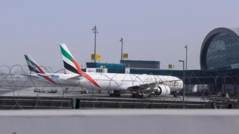 Reuters Two Emirates planes parked at Dubai Airport. They are both white with green, red, white and black stripes on its tail.