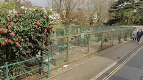 Google General view of a bird aviary. The aviary is a long rectangular-shaped structure made of wooden posts and fencing. A number of birds are inside the aviary. It is next to a pavement and road. Pedestrians are walking along the pavement and are looking at the birds. 