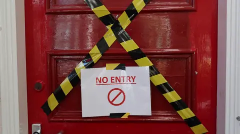 Hope Rhodes / BBC A red door has yellow warning tape across it in the shape of an X. In the middle of the door a laminated A4 sign reads no entry in red writing.