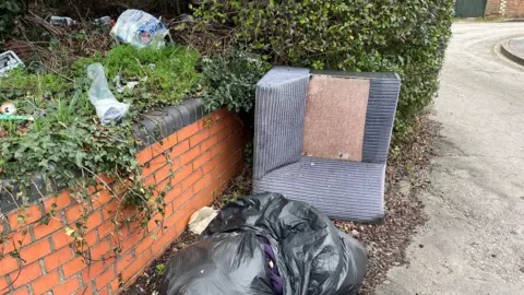 An upturned armchair, two full black bin bags are on the ground, next to a small brick wall on a residential back road. Plastic bags, empty cans and plastic bottles are littered on grass above the wall. 