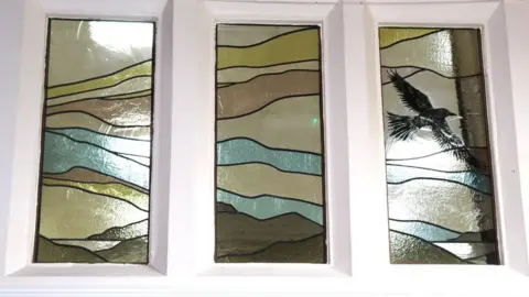 J and P R A close up view of three panels of stained glass showing a series of wavy lines and a black bird in flight