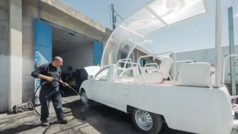 A man with a power hose works on the popemobile at a garage.