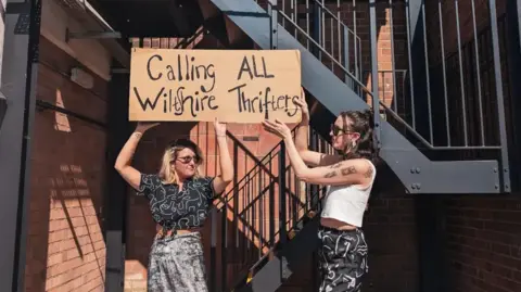 Swap Society Two women stood by an outdoor staircase in stylish clothes. They hold a cardboard sign which says 'Calling All Wiltshire Thrifters'