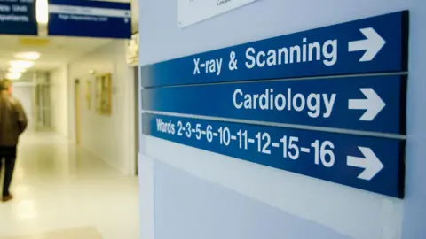 Getty Images A stock image of a hospital sign pointing towards cardiology. It is a blue sign with white font. Above it a sign reads "X-ray & Scanning" while below it is a sign pointing to wards numbered from 2 to 16.