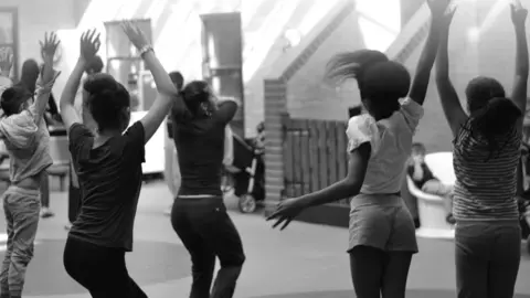 Black Country Women's Aid Several women and girls have their hands in the air with their backs to the camera. They look to have been dancing as they stand in an open area with a brick staircase at the far end of the room. The women wears dark trousers and tops, one of the girls has shorts and a white top, they all have dark hair.