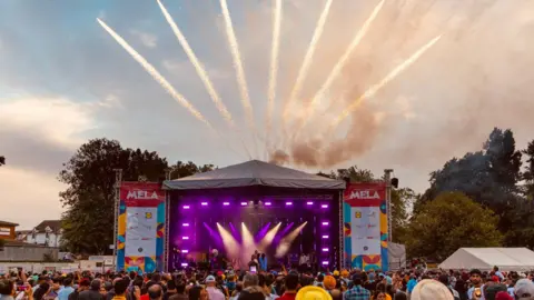 A photo of a stage with purple lighting and crowds of people stood in front of it. Pop-up signs near the stage say 'MELA'. There are sparks of light rising from above the stage.