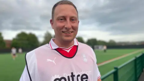 Simon Thake A man with closely cropped hair smiles next to a football pitch. He wears a white bib over a red football shirt.
