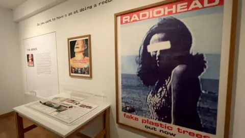 The album cover for Radiohead's The Bends is framed and hanging on the wall of an art gallery. Alongside it is a much larger framed poster for the single Fake Plastic Trees which features a photograph of woman who's identity has been hidden by a white strip across her eyes. There's a collection of items in a glass case on a table below the artwork.