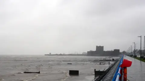 A stormy seaside promenade with rough waves, grey sky, and a distant castle in the rain.