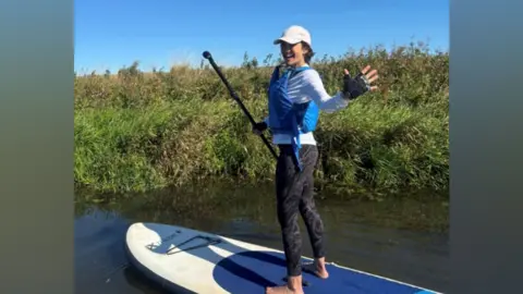 Myeloma UK A woman wearing a blue life jacket and sports gear. She is paddle boarding.