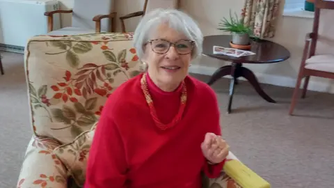 Gail Ratcliffe A woman sits in an armchair and smiles at the camera. She has short grey hair and wears a red jumper with a red and yellow necklace. 